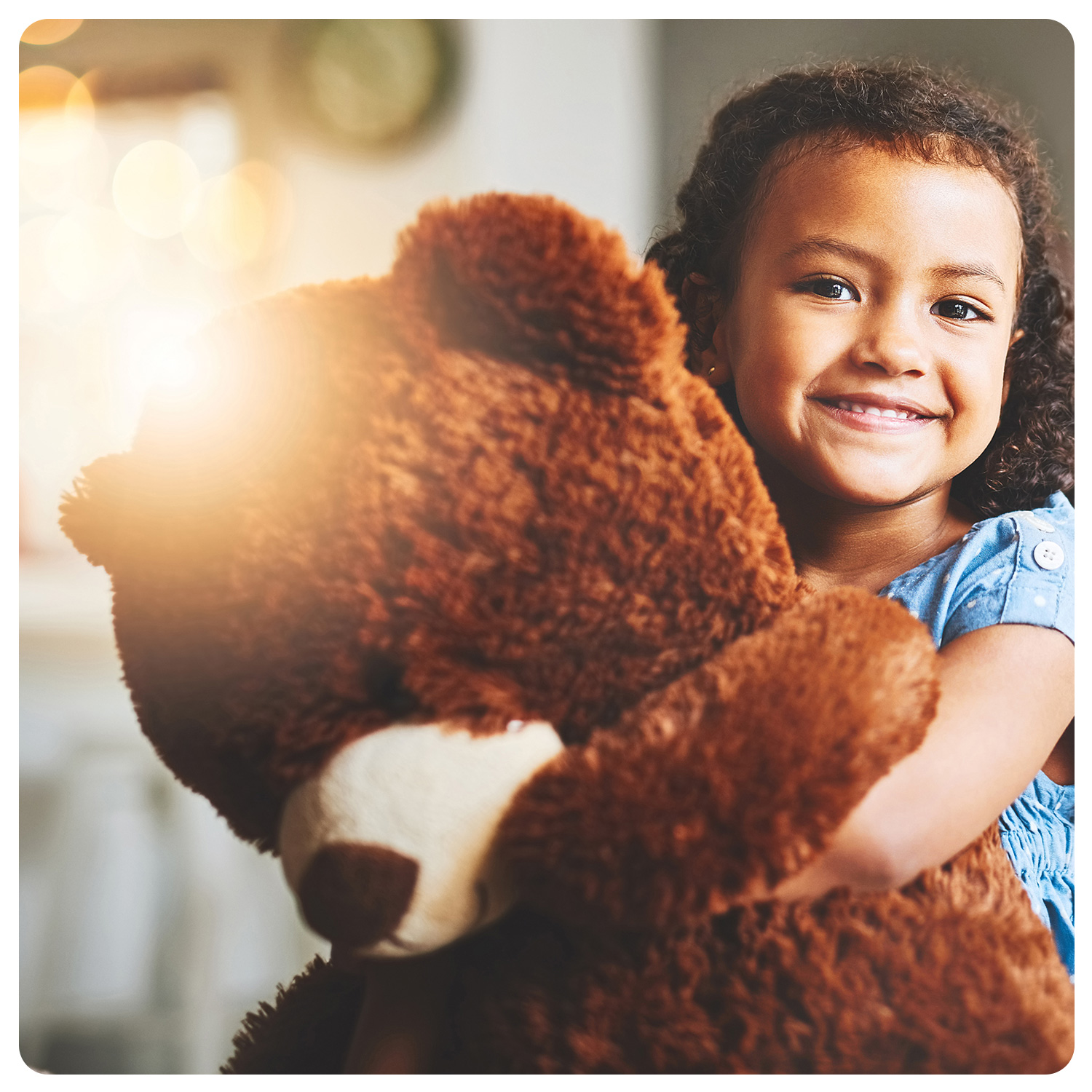 Ein junges Mädchen mit lockigem Haar lächelt, während es einen großen braunen Teddybären umarmt. Sie trägt ein hellblaues Kleid, während im Hintergrund warmes Sonnenlicht scheint, das eine fröhliche und familiäre Atmosphäre einfängt.